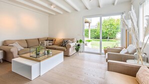 Un salon moderne avec un canapé beige, une table basse en verre et une vue sur un jardin, visible à travers une porte-fenêtre coulissante.