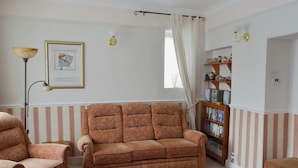 Uma sala de estar com um sofá estampado, uma cadeira, um abajur, uma estante de livros e um quadro na parede.