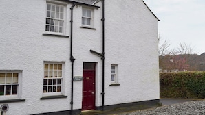 Um edifício branco com porta vermelha, uma placa com a inscrição 'Daisy Bank Cottage' e um muro de pedra ao fundo.