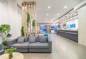 A modern reception area with a grey sofa, wooden vertical panels with greenery, a reception desk, and a tiled floor.