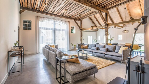 Un'ampia zona giorno con soffitto alto e travi in legno a vista, caratterizzata da un grande divano componibile, un tavolino in vetro e una lampada moderna.