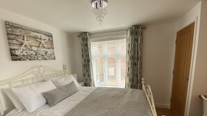 A neatly made bed with a textured blanket, a wall-mounted artwork of seashells, and a window with patterned curtains. - Perfect for beach & nature reserve walks all year round (Pagham)