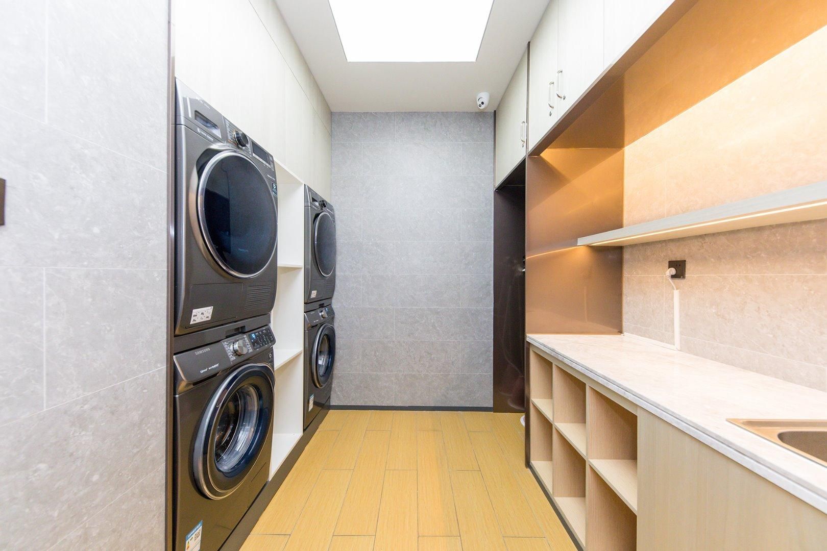 A compact laundry room with two front-loading washing machines, a countertop with a sink, and a skylight above.