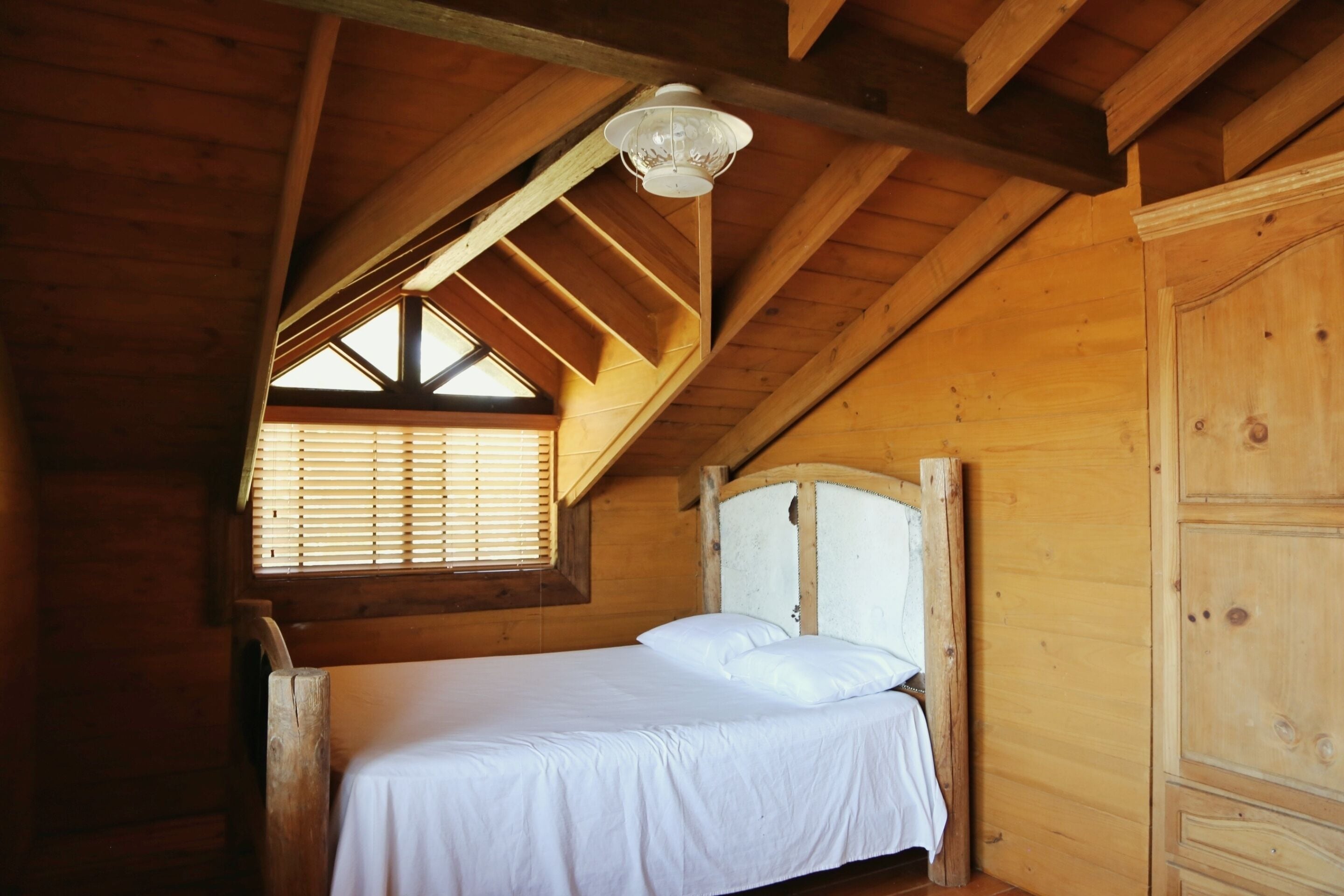 A wooden cabin room with a bed, a window with blinds, and a wooden wardrobe.