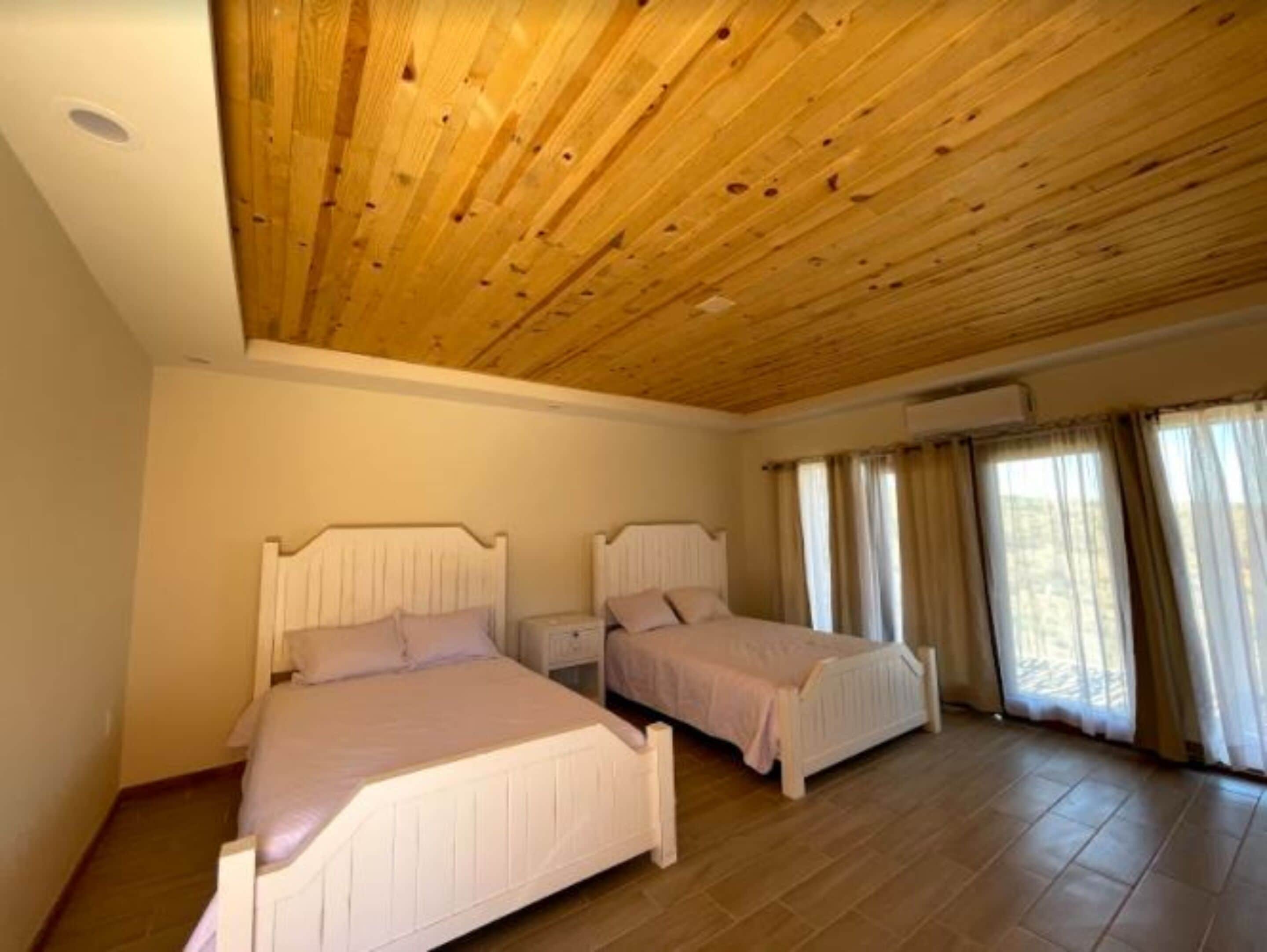 Une chambre avec deux lits, un plafond en bois et de grandes fenêtres avec des rideaux.