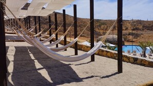 Varias hamacas bajo una zona sombreada, con una piscina y sillones de descanso al fondo.