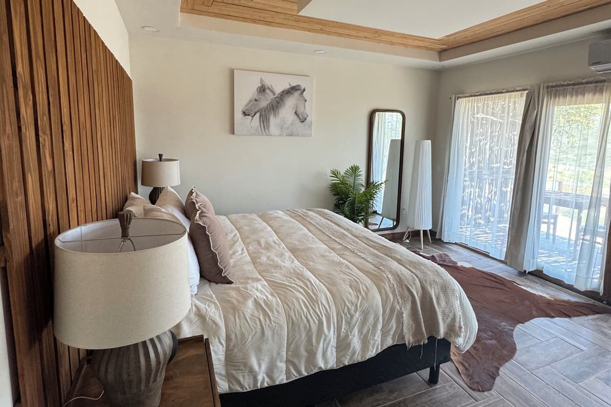 A bedroom with a wooden ceiling, a large bed with a white bedspread, a bedside lamp, and a painting of horses on the wall.