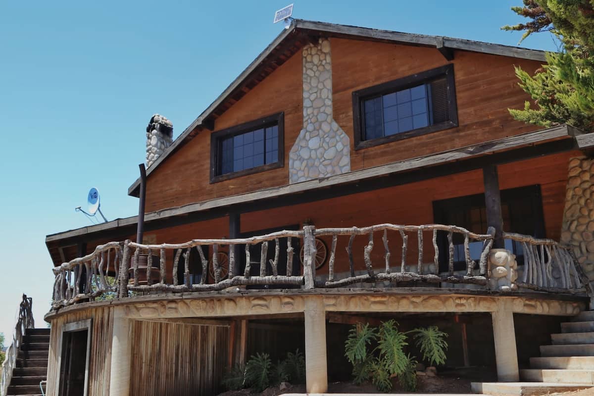 A two-story wooden cabin with a stone chimney, a balcony with a rustic railing, and a satellite dish.
