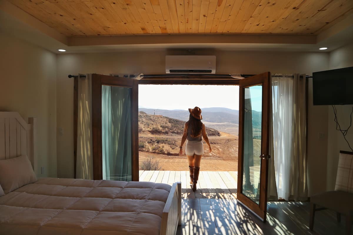 A bedroom with a large bed, a wooden ceiling, and a view of a desert landscape through an open door.