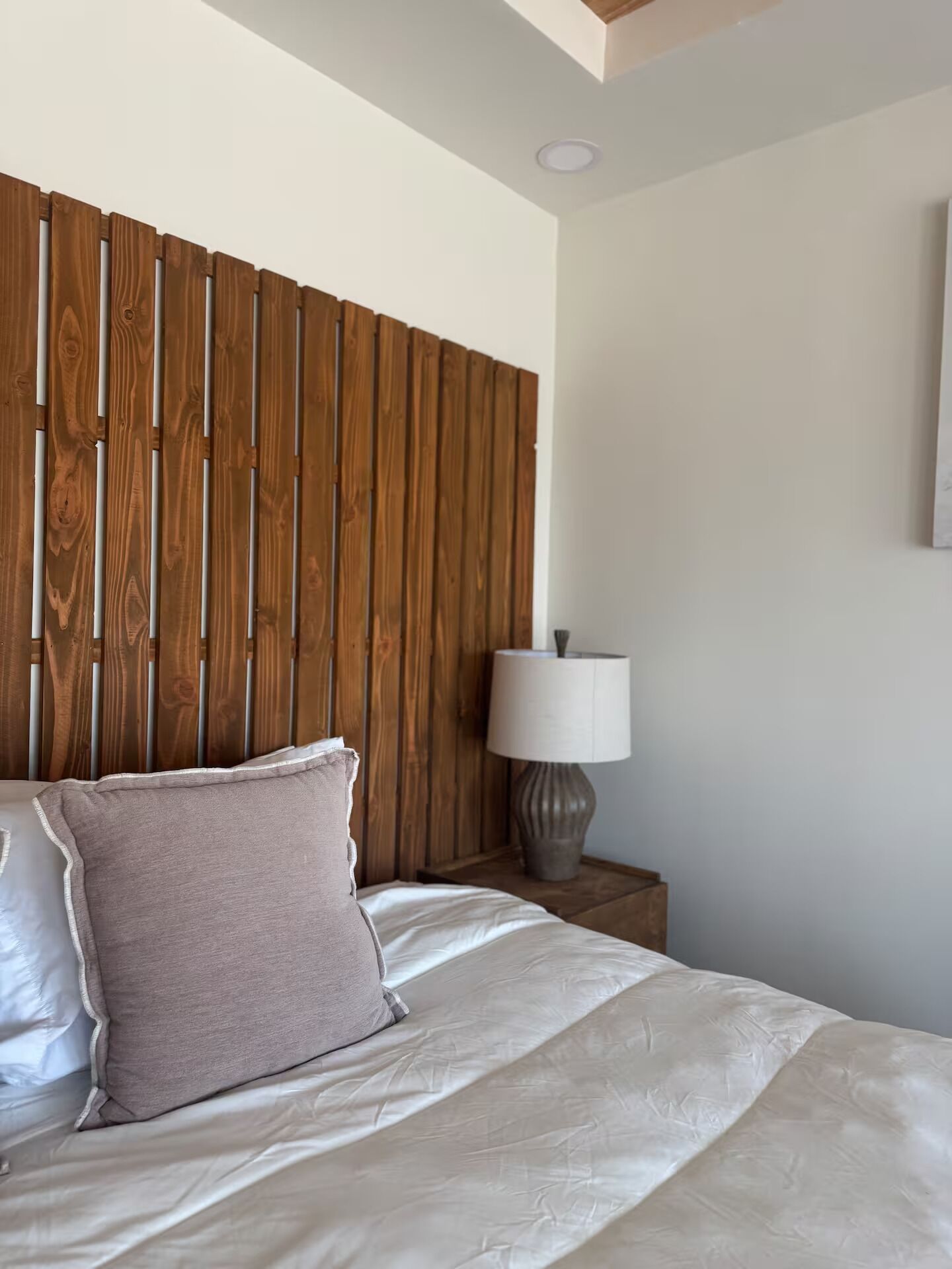 A bedroom with a wooden headboard, a bed with white bedding, a nightstand with a lamp, and a wall-mounted light fixture.