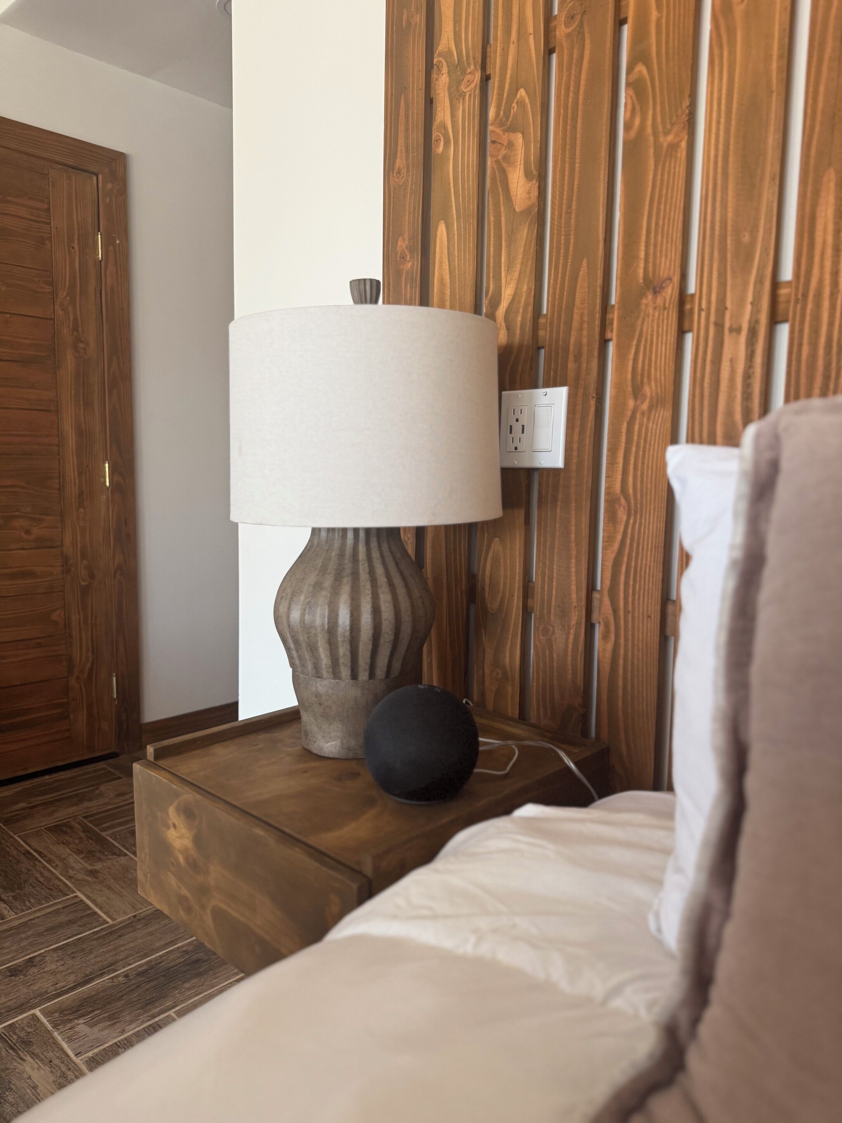 Una mesita de noche de madera con una lámpara y un objeto negro, posiblemente un altavoz, al lado de una cama con un almohadón blanco.