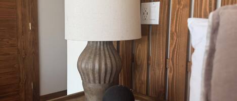 Una mesita de noche de madera con una lámpara y un objeto negro, posiblemente un altavoz, al lado de una cama con un almohadón blanco.