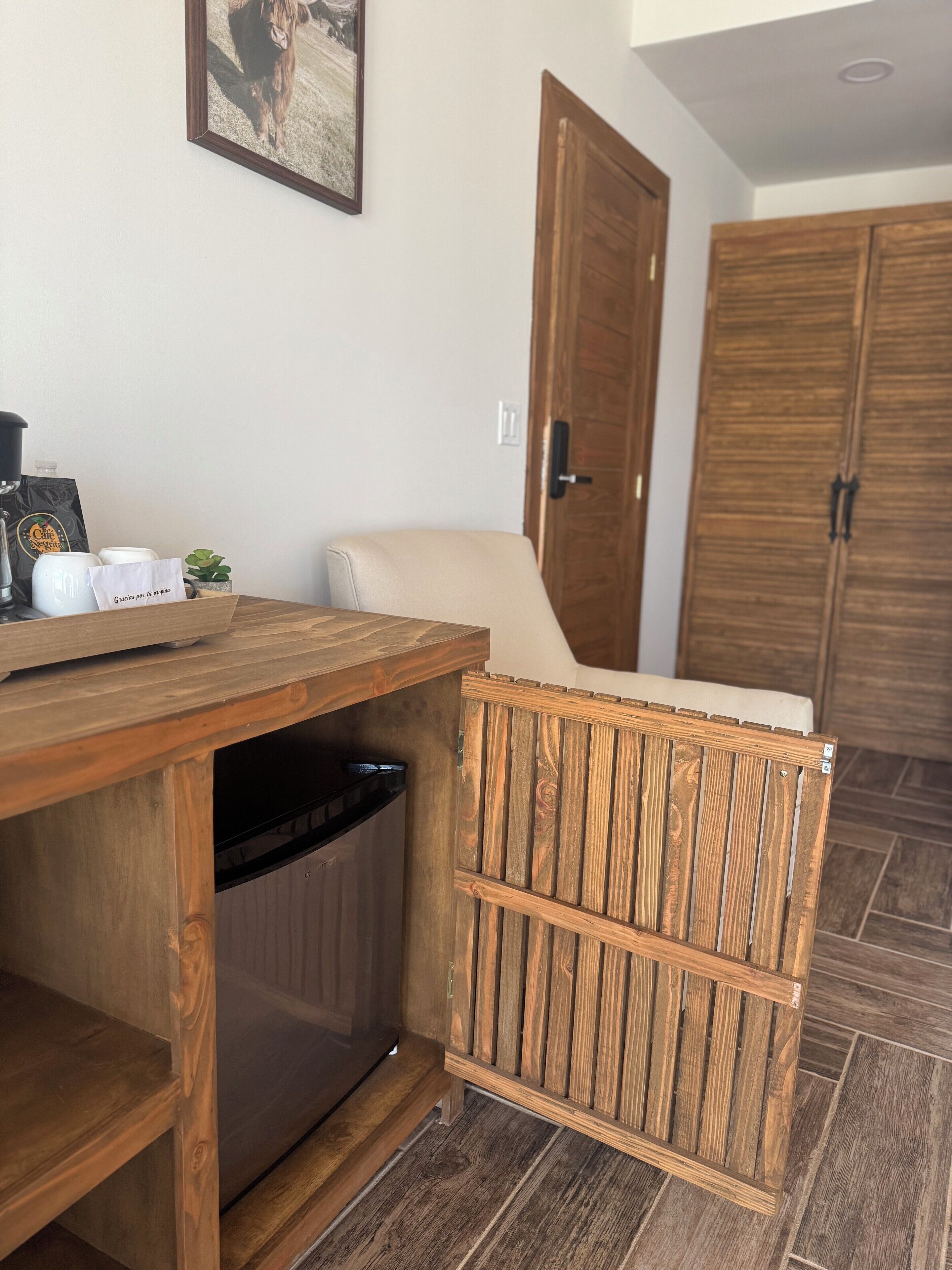 Escritorio de madera con un cubo de basura, una planta pequeña y una máquina de café. Se observa una silla de madera con cojín y una puerta de madera al fondo.
