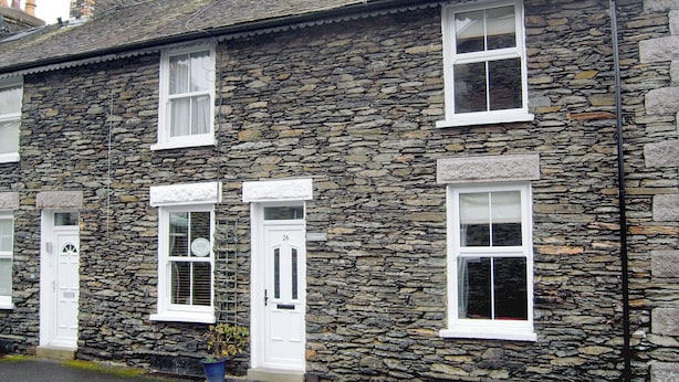 A stone-built two-story house with white-framed windows and doors, a visible number '26' on the door, and a small potted plant at the entrance.