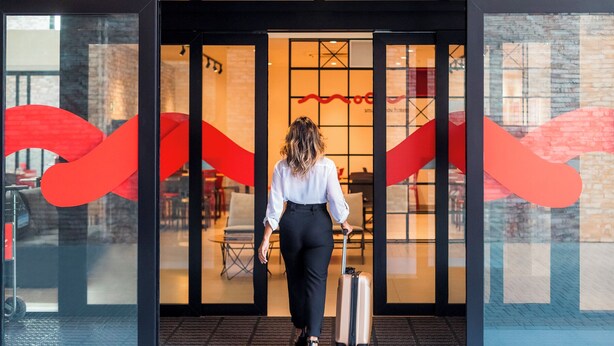 A person with a suitcase entering a building through a glass door with a red abstract design.