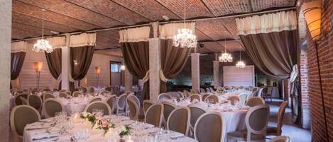Uma sala de banquetes com mesas redondas preparadas para refeições, cortinas decorativas e lustres.