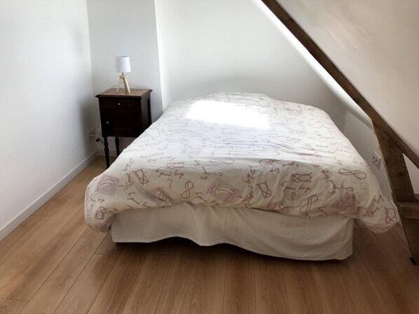 Une chambre avec un plafond en pente, un lit recouvert d’une couvre-lit à motifs, une table de chevet avec une lampe et un plancher en bois.