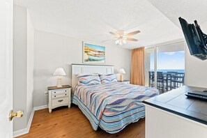 A bedroom with a bed, nightstands, a desk, a ceiling fan, a painting, and a view of the ocean.