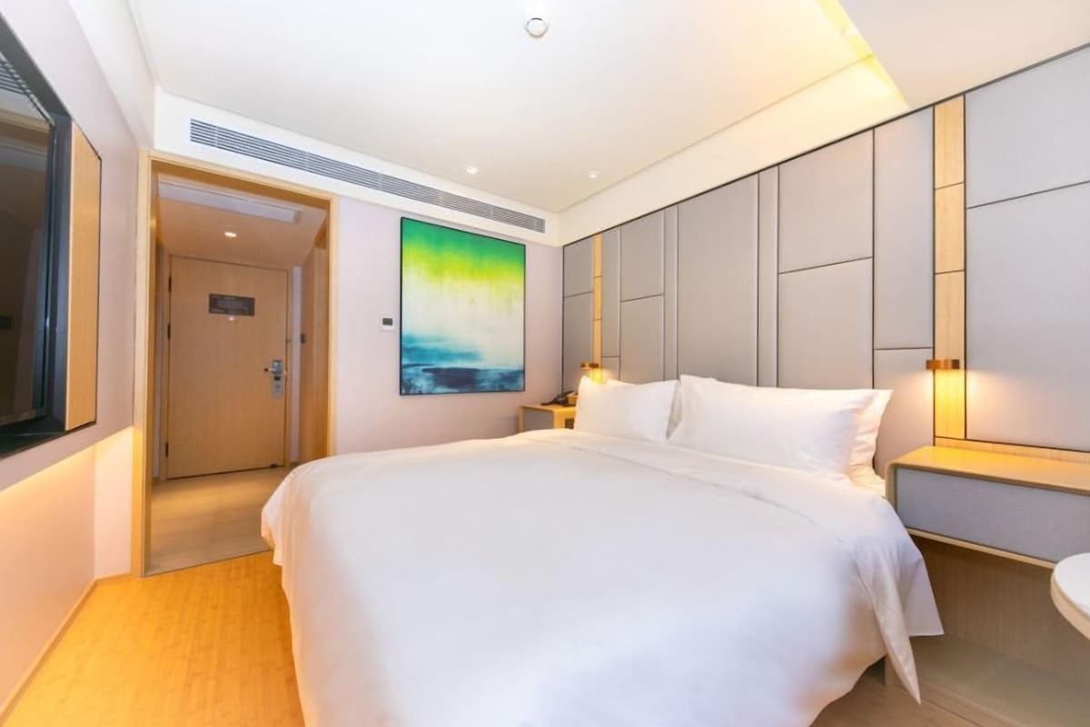 A hotel room with a large bed, a painting on the wall, a bedside table with a lamp, and a door leading to another room.