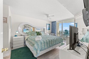 Un dormitorio con cama, escritorio con computadora, ventilador de techo y ventana grande con vistas.