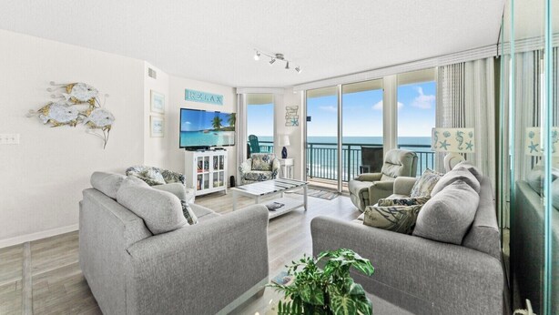 A spacious living room with a large TV, comfortable seating, a balcony with a view of the ocean, and a 'RELAX' sign on the wall. - 4-bedroom, 3-bathroom, family-friendly oceanfront condo, 9th floor views | Windy Hill Dunes - 906 (North Myrtle Beach)