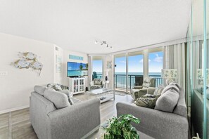 A spacious living room with a large TV, comfortable seating, a balcony with a view of the ocean, and a 'RELAX' sign on the wall.