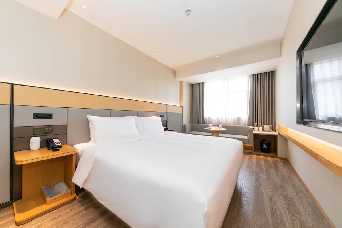 A hotel room with a large bed, a bedside table with a phone and cup, a small table with a lamp, a sofa, and a window with curtains.