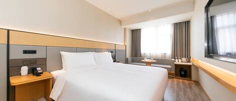 A hotel room with a large bed, a bedside table with a phone and cup, a small table with a lamp, a sofa, and a window with curtains.