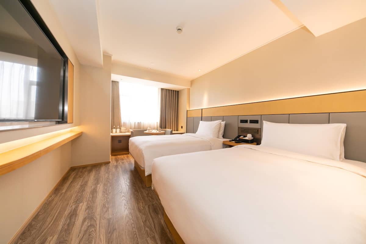 A hotel room with two beds, a wooden floor, a large window with curtains, a flat-screen TV, and a bedside table with a lamp.