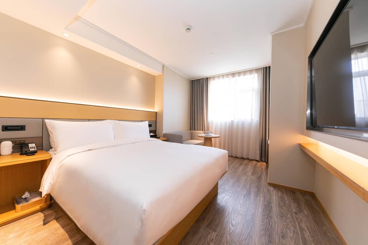 A hotel room with a large bed, bedside tables, a desk, a chair, a TV, and a window with curtains.