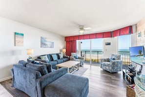 A living room with a sofa, chairs, a coffee table, a TV, and a balcony with a view of the ocean.