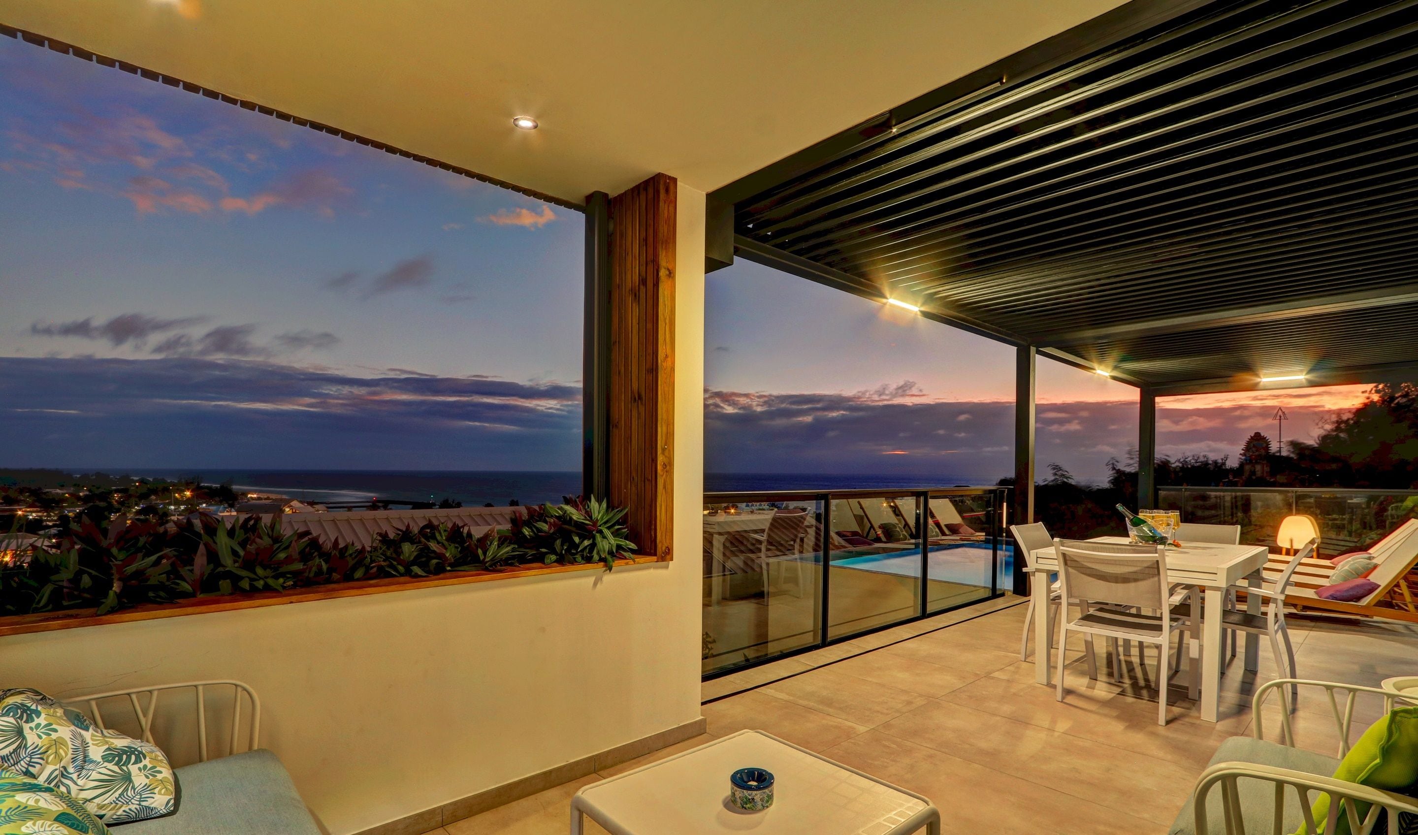 A balcony with a view of the sea, a pool, and a dining area with chairs and a table.