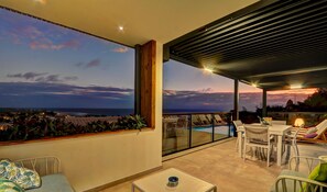 A balcony with a view of the sea, a pool, and a dining area with chairs and a table.