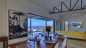 A dining area with a table set for two, a large painting of tropical foliage, and a view of the ocean. - La Villa Azur Saint-Gilles ***** - Reunion Island - Seaview, Pool, Beach & BBQ! (Saint-Gilles les Bains)
