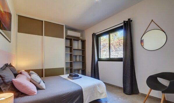 A modern bedroom with a bed, a built-in wardrobe, a small table with a lamp, a chair, and a window with curtains.