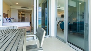 A modern hotel room with a bed, a wooden table, and a bench. There is a sliding glass door leading to a balcony with a blue sofa and a dining area.