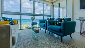 A modern living room with a large window overlooking the ocean, two teal armchairs, a gray sofa, and a glass coffee table.