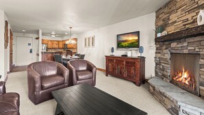 Una sala de estar con sillones de cuero, una mesa de centro de madera oscura, una chimenea de piedra, una cocina al fondo y un televisor montado en la pared.