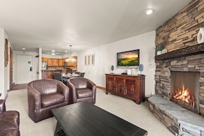 A living room with leather chairs, a dark wooden coffee table, a stone fireplace, a kitchen in the background, and a mounted television.