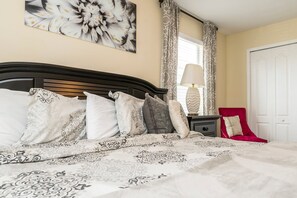 A neatly made bed with patterned bedding, a bedside table with a lamp, and a red chair. - Paradise Palms 2958 (Kissimmee)