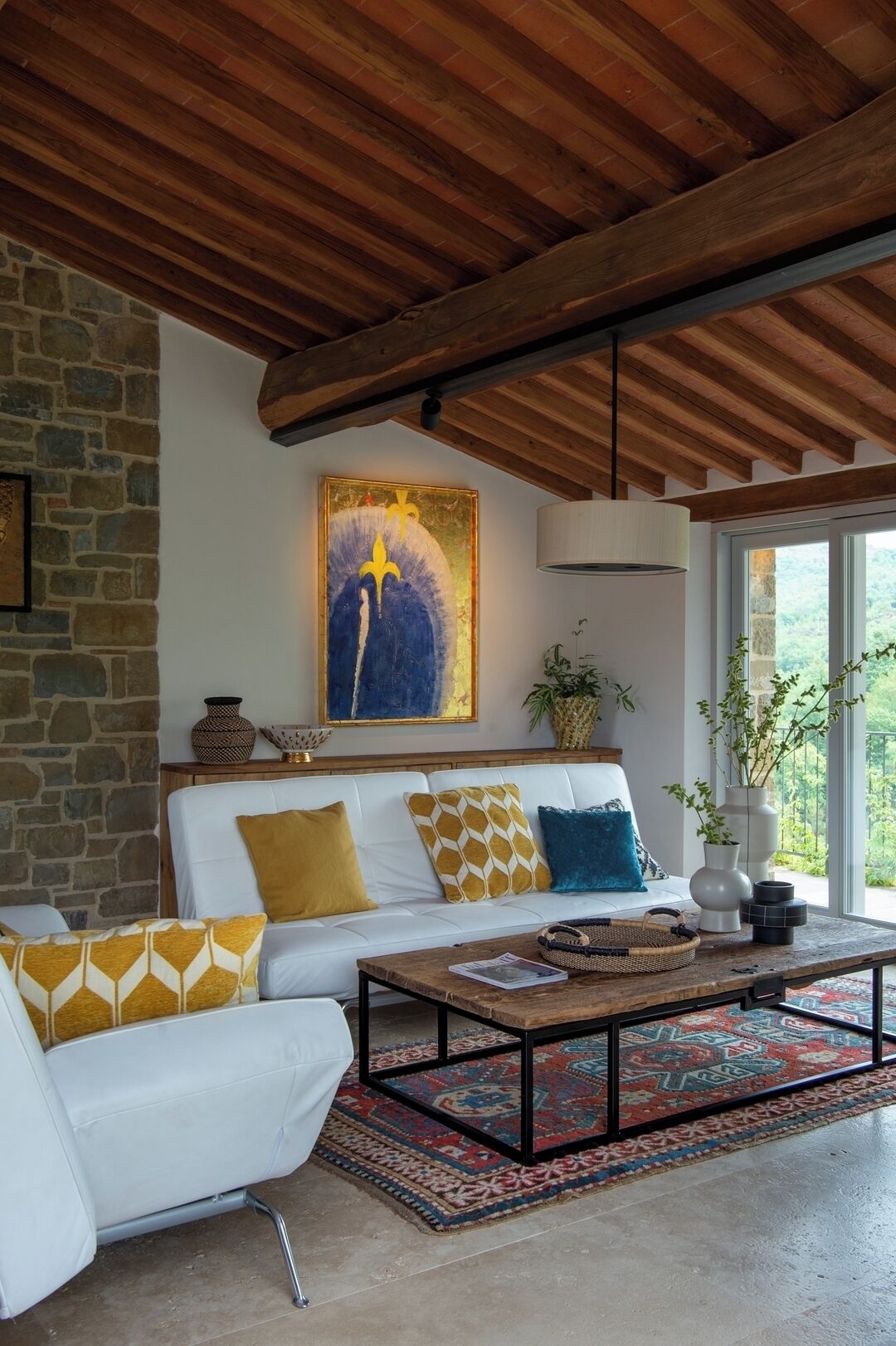 A modern living room with a stone wall, a white sofa with patterned cushions, a wooden ceiling, and a large window with a view of greenery.