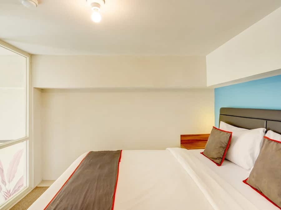 Una habitación de hotel con un ventanal grande, una cama con ropa de cama blanca y detalles rojos, y una pared de acento azul.