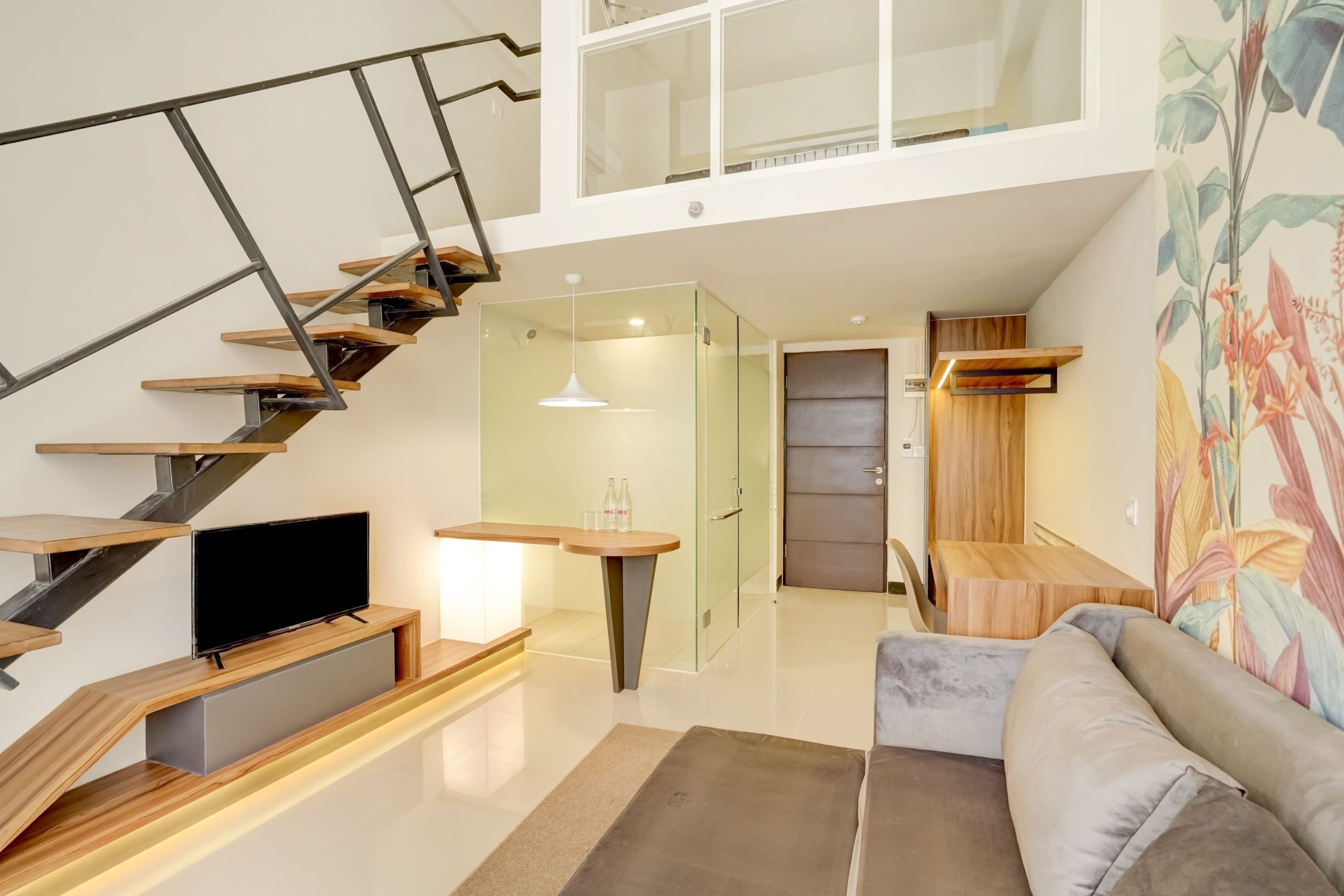A modern living space with a staircase, a flat-screen TV, a sofa, and a glass door leading to another room.