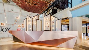 Balcão de recepção moderno com design geométrico e detalhes de borboleta, situado em um hall amplo com pé direito alto e detalhes em madeira.
