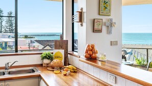 A kitchen with a view of the sea, a wooden countertop, a bread loaf, a glass of orange juice, and a bowl of oranges. - Namanula Unit 6 - Ocean Views & Walk to Convent Beach - Holiday Yamba (Yamba)