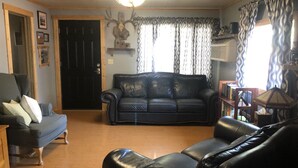 Uma sala de estar com um sofá de couro preto, uma poltrona cinza e uma mesa de centro de madeira. Há uma porta preta com painel decorativo, um candelabro e uma janela com cortinas estampadas.
