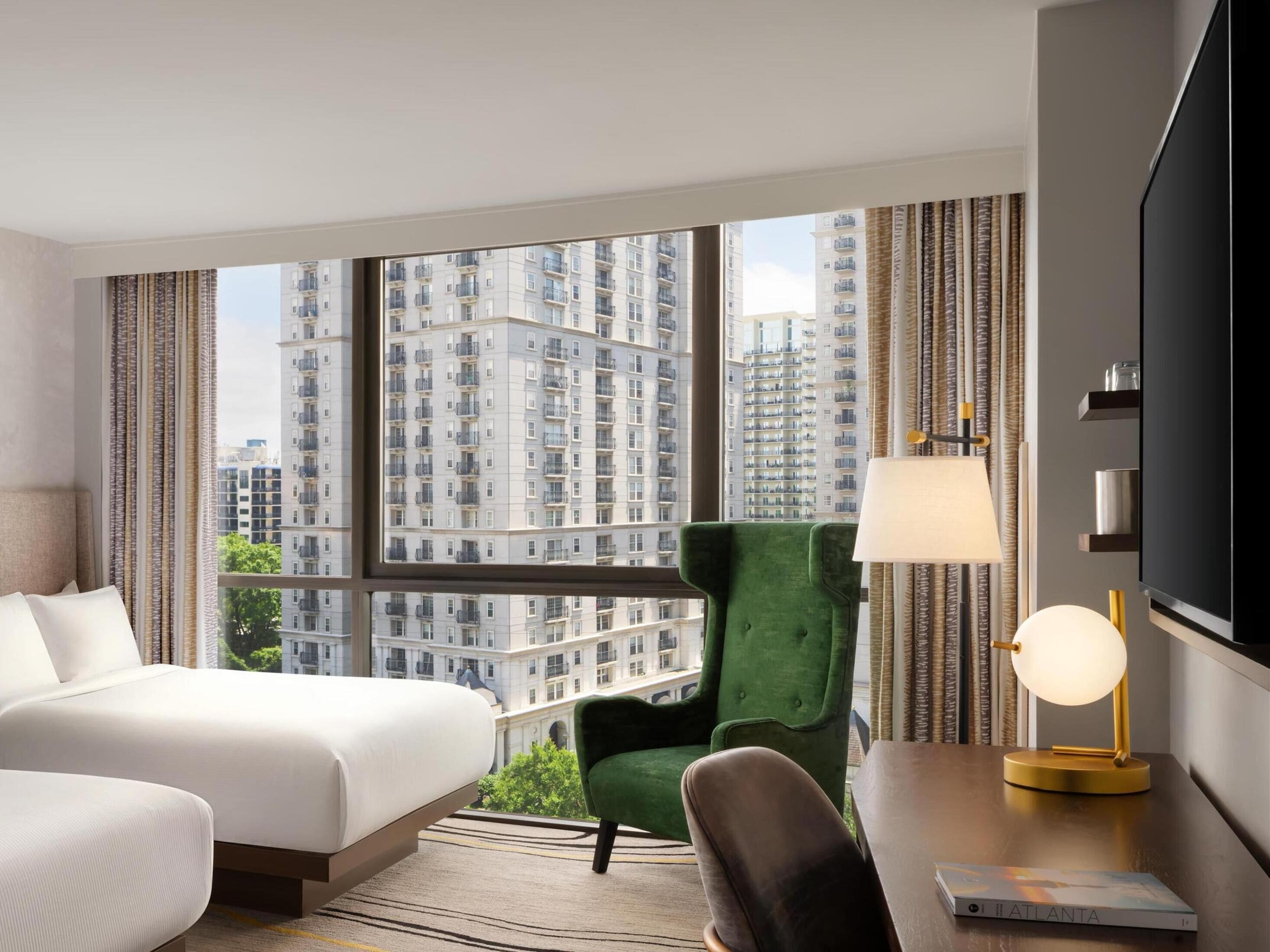 A hotel room with a bed, a green armchair, a desk with a lamp, and a TV.