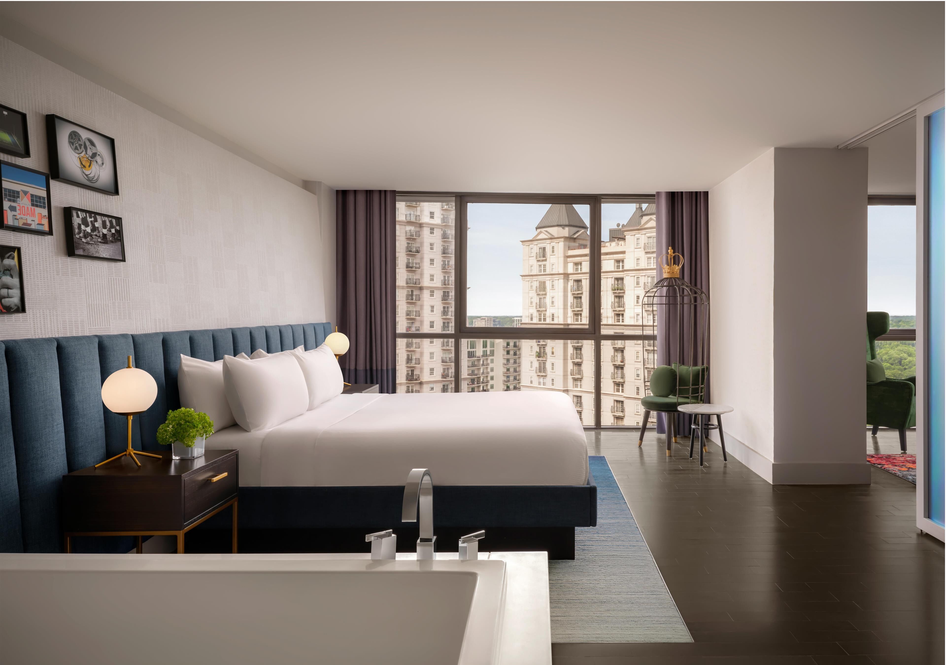 A modern hotel room with a large bed, a bedside table with a lamp, a bathroom with a sink, and a view of high-rise buildings.