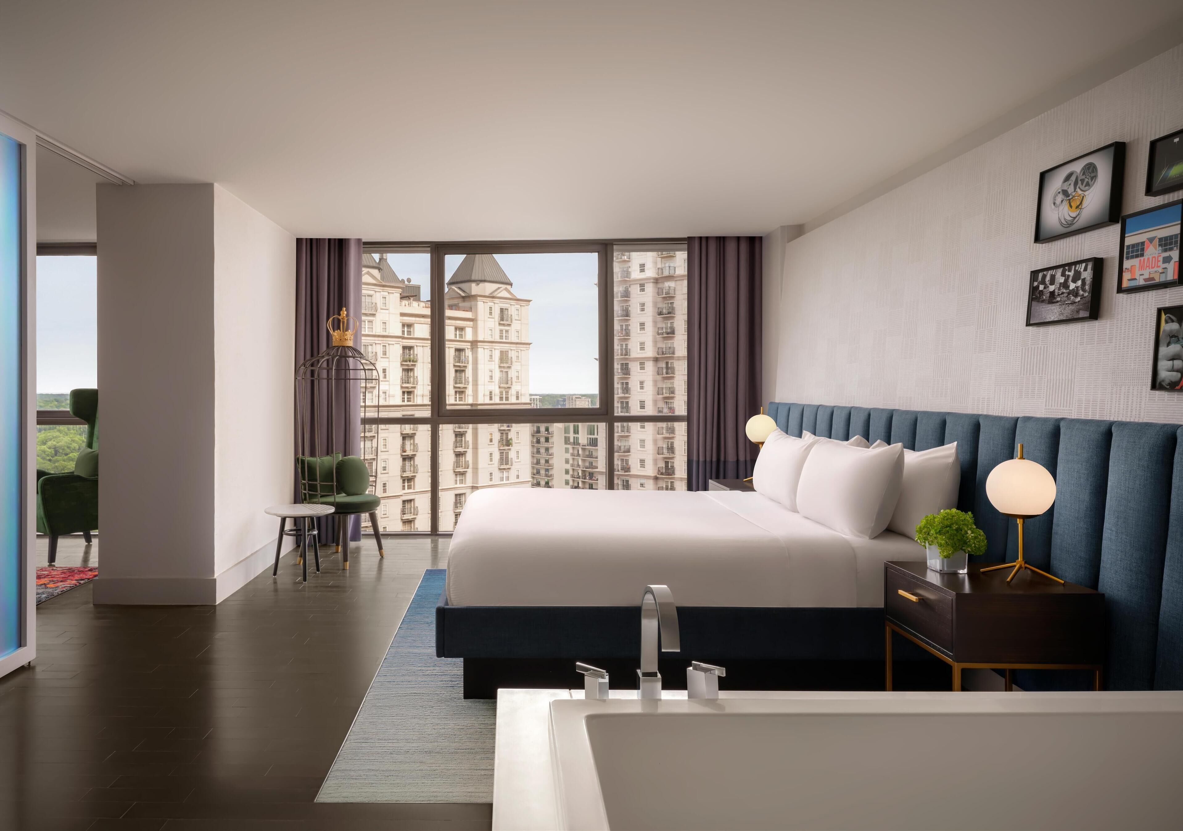 A modern hotel room with a large bed, a bedside table with a lamp, a view of high-rise buildings, and a small seating area with a chair and table.