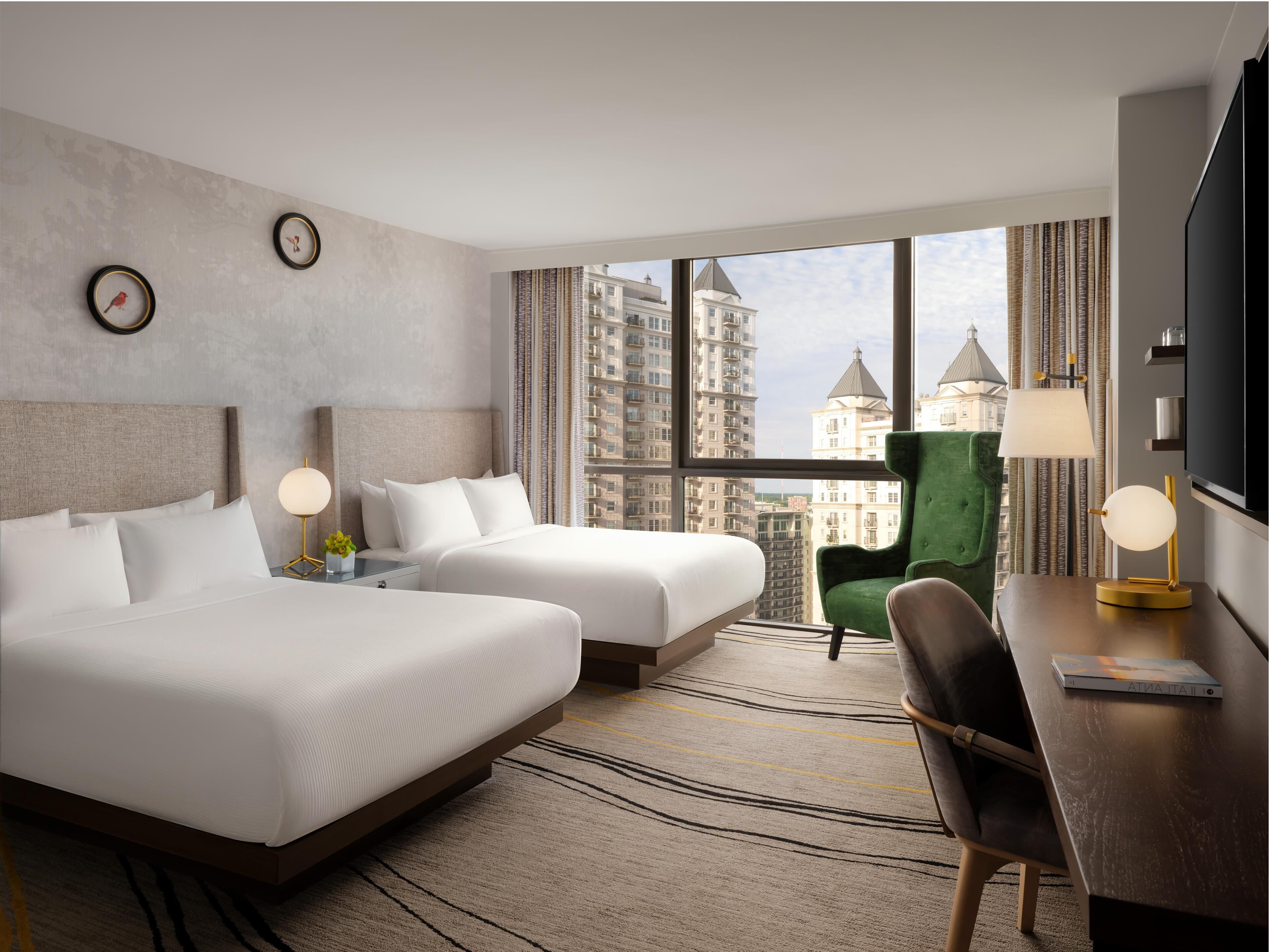 A hotel room with two beds, a green armchair, a desk with a lamp, and a TV.